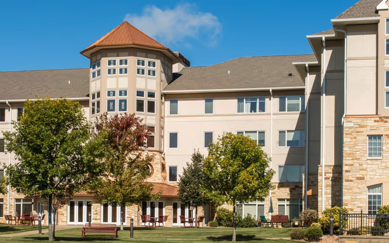 Exterior view of a senior living facility building with multiple windows, a turret-like architectural feature, and a mix of beige and stone facade. There are several trees and benches on a well-maintained lawn in front of the building under a clear blue sky.
