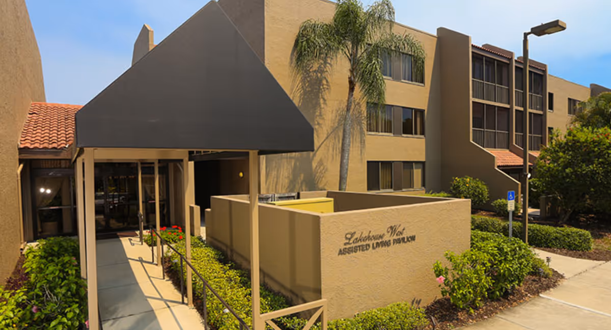 Exterior view of Lakehouse West Assisted Living Pavilion showing the entrance with a covered walkway, beige building walls, windows, palm trees, and landscaped greenery along the pathway.