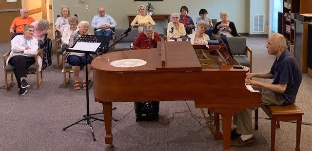 A group of elderly people seated in chairs and wheelchairs in a common room, attentively listening to a man playing a grand piano. The room has a carpeted floor and light-colored walls with a door and some furniture in the background.