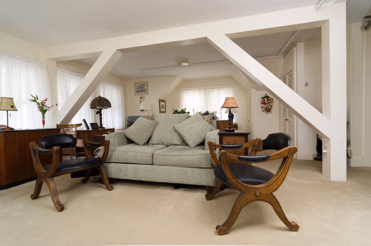 Bright living room with a light-gray sofa, two wooden accent chairs, side tables and exposed white beams.