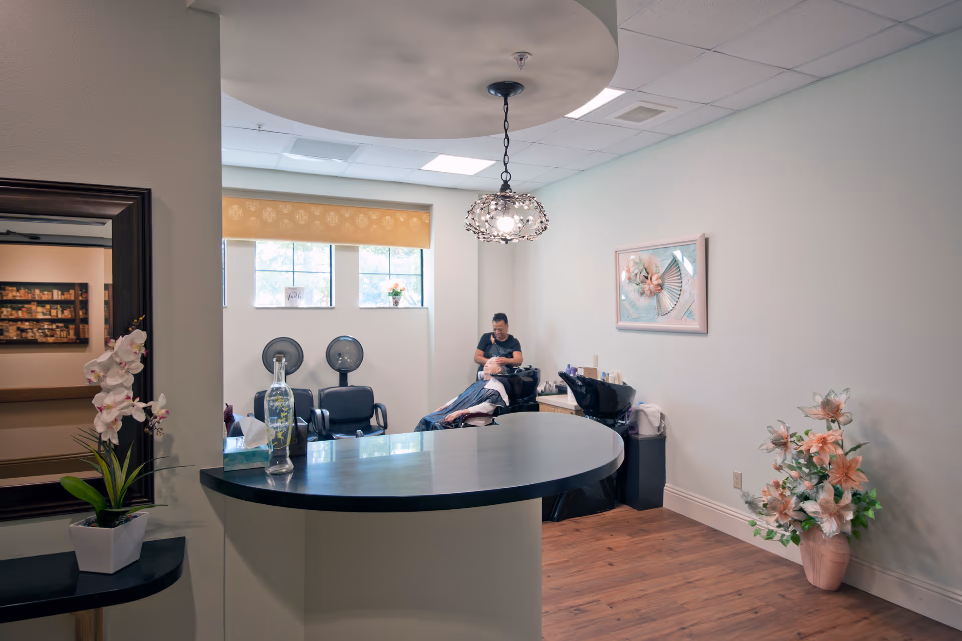 Interior of a salon area in a senior living facility with a stylist washing a resident's hair at a shampoo station. The room has a curved counter in the foreground, two black salon chairs with hair dryers near the window, a decorative hanging light fixture, a framed floral artwork on the wall, and a large flower arrangement in a vase on the floor.