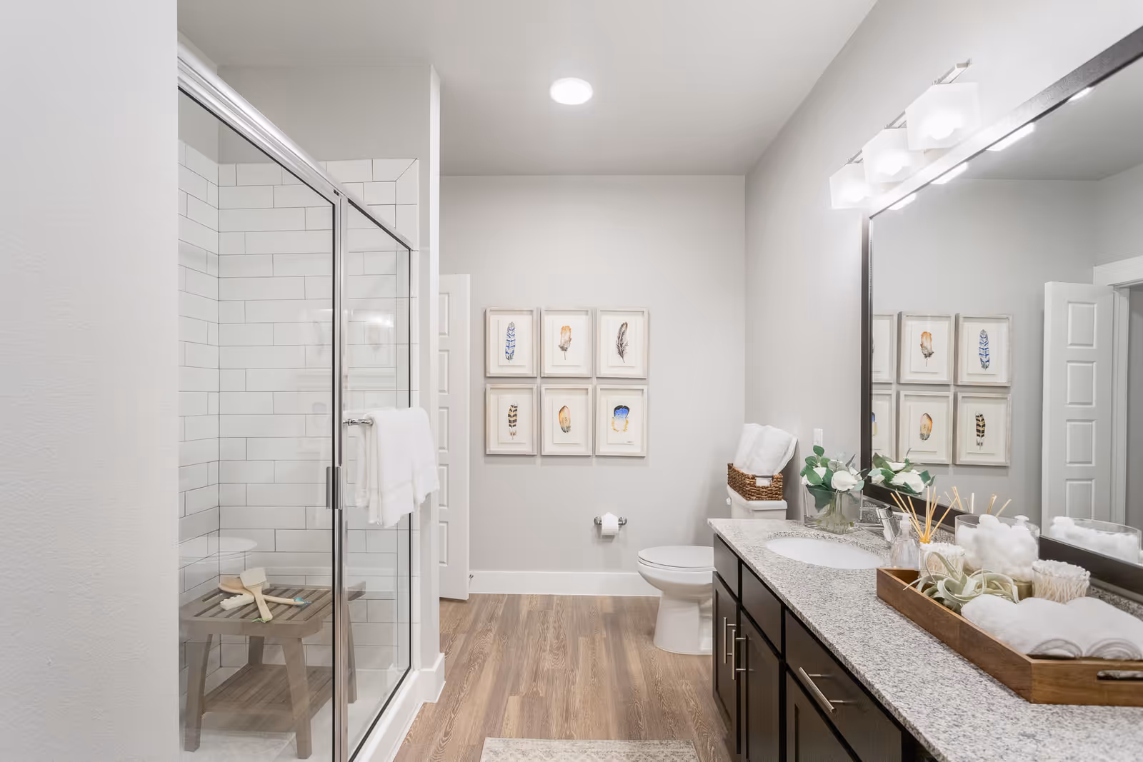 A modern bathroom featuring a glass shower, a double sink vanity with granite countertops, decorative wall art, and wooden flooring.