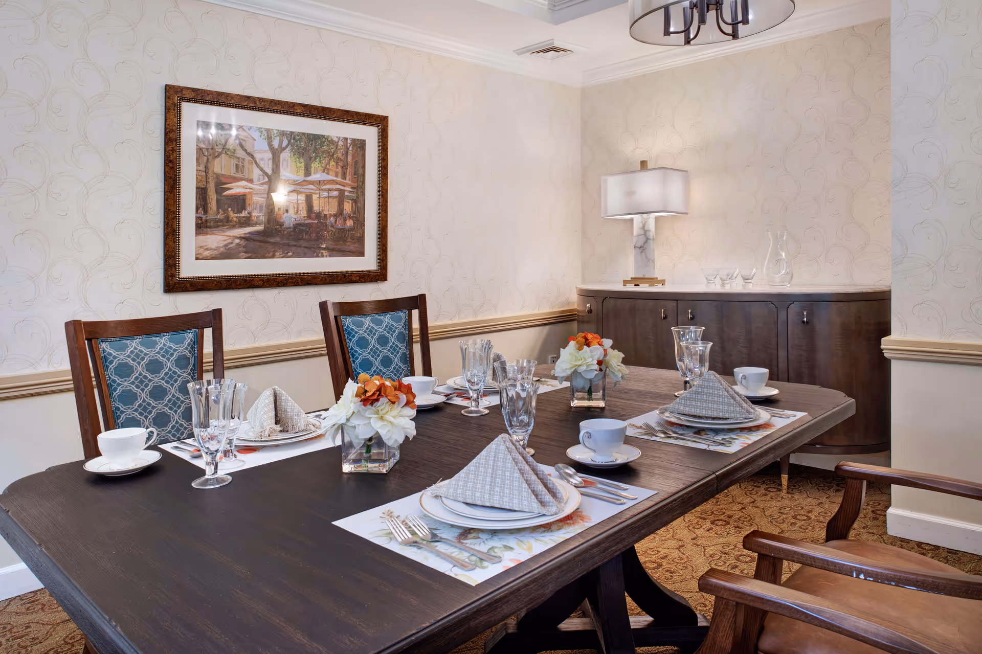 A dining room with a dark wooden table set for four people, featuring folded napkins, plates, cups, glasses, and floral centerpieces. The room has patterned wallpaper, a framed painting on the wall, a sideboard with a lamp and glassware, and upholstered wooden chairs.