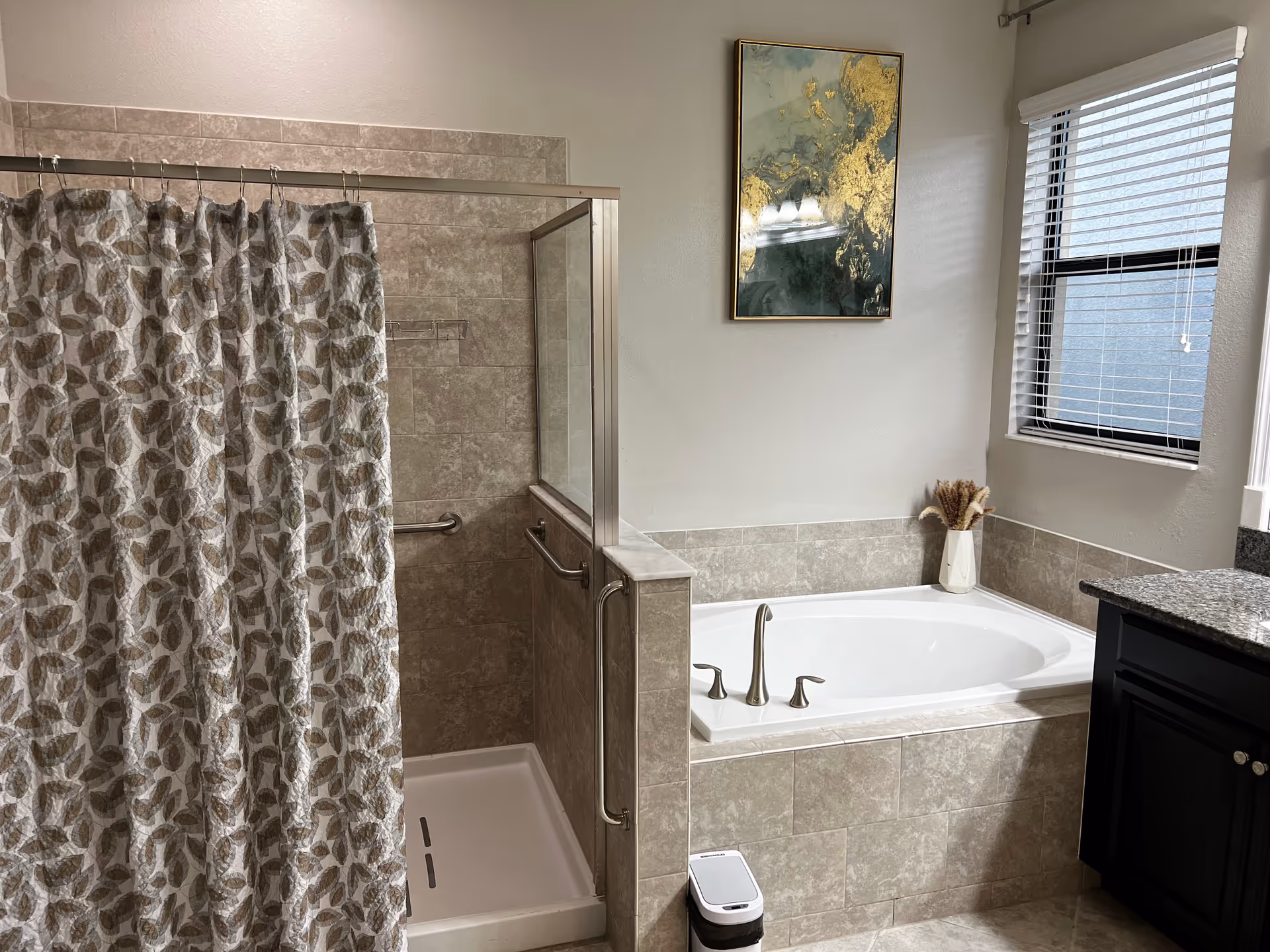 A bathroom featuring a tiled walk-in shower with a leaf-patterned shower curtain, a built-in bathtub with silver faucets, a window with blinds, a dark wood vanity with a granite countertop, a small trash can, and a decorative vase with dried plants. A framed abstract painting hangs on the wall above the bathtub.