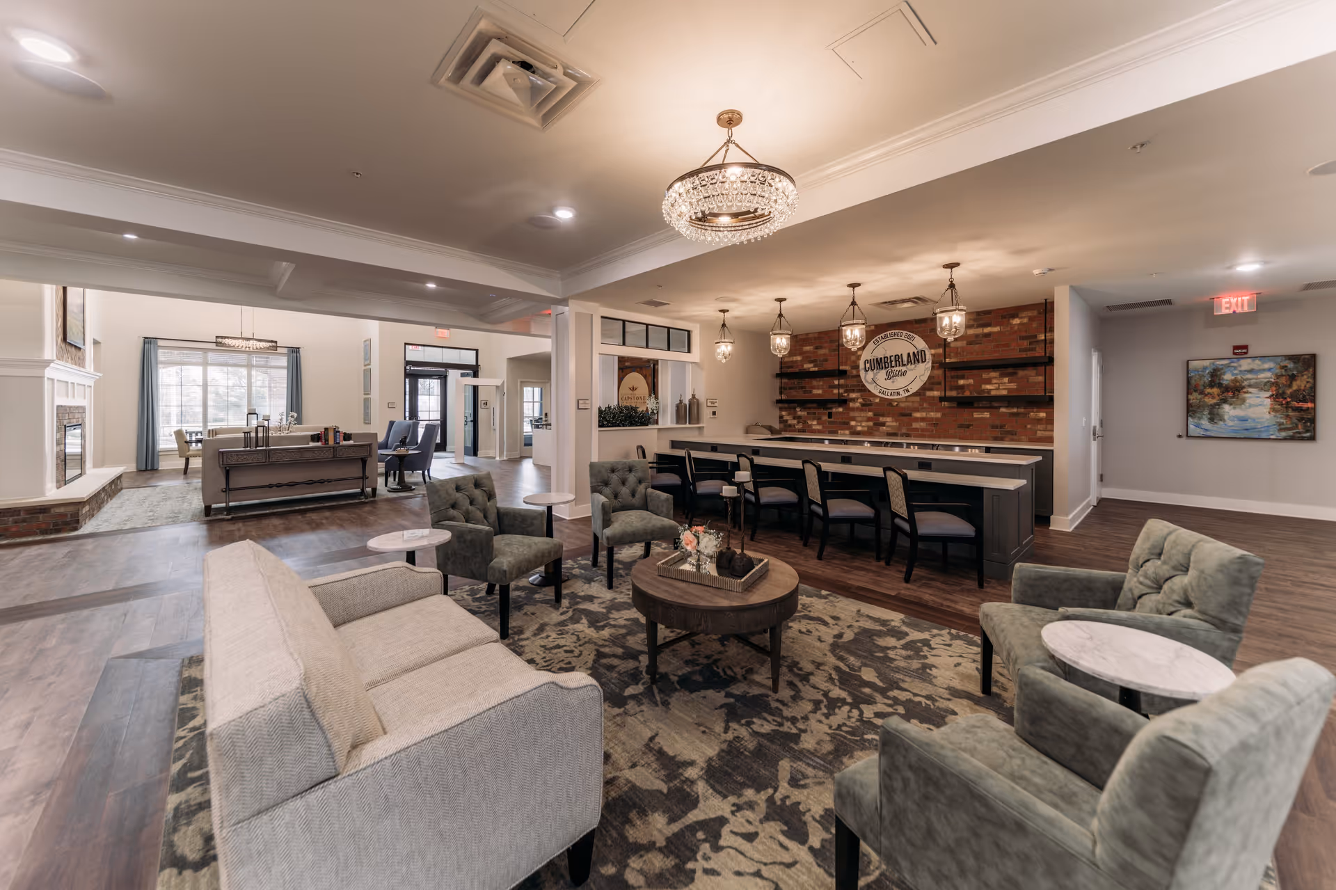 A spacious and well-lit common area in a senior living facility featuring a seating arrangement with a beige sofa, four green armchairs, and round side tables on a patterned rug. In the background, there is a bar area with high chairs and pendant lights hanging from the ceiling. The walls have a mix of brick and light paint, with decorative elements including a sign that reads 'Cumberland Station' and a framed painting. Large windows and a fireplace are visible in an adjacent room.