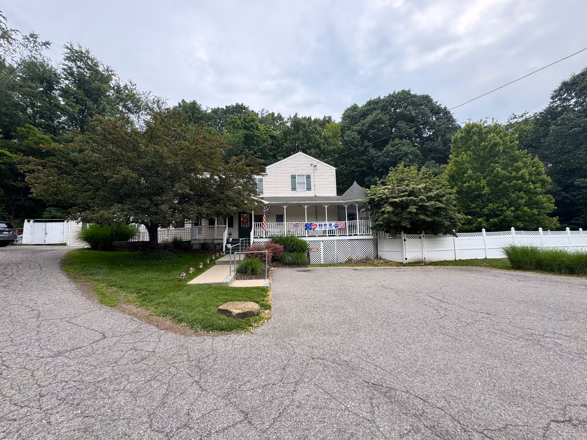 Exterior view of a two-story white building with a wrap-around porch decorated with patriotic banners and an American flag. The building is surrounded by trees and a white fence, with a paved driveway and parking area in front.