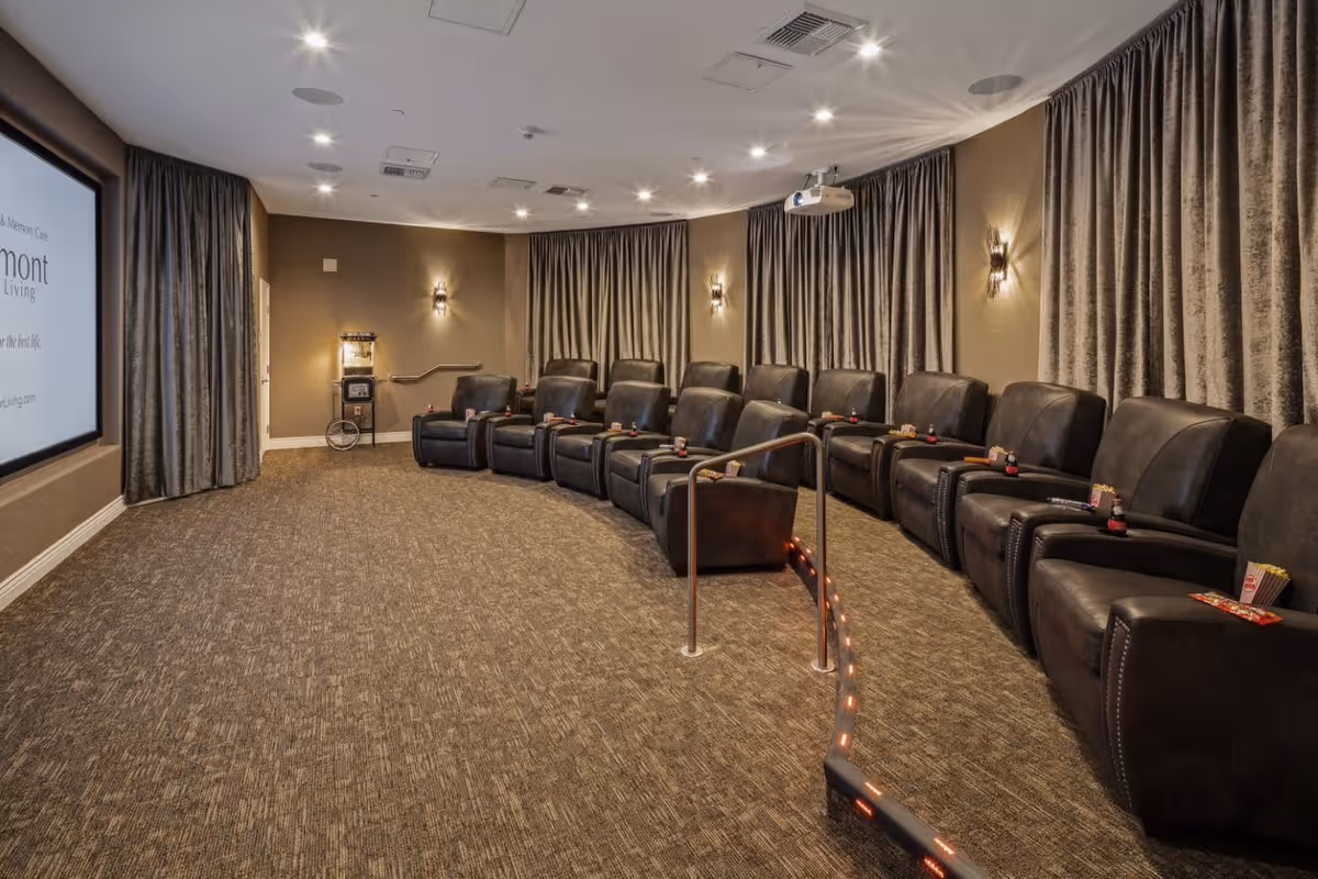 A cozy private movie theater room with two rows of black leather recliner chairs arranged in a curved formation facing a large screen. The room has carpeted flooring, beige walls, wall sconces for lighting, and heavy curtains covering the windows. A popcorn machine is visible in the back corner.