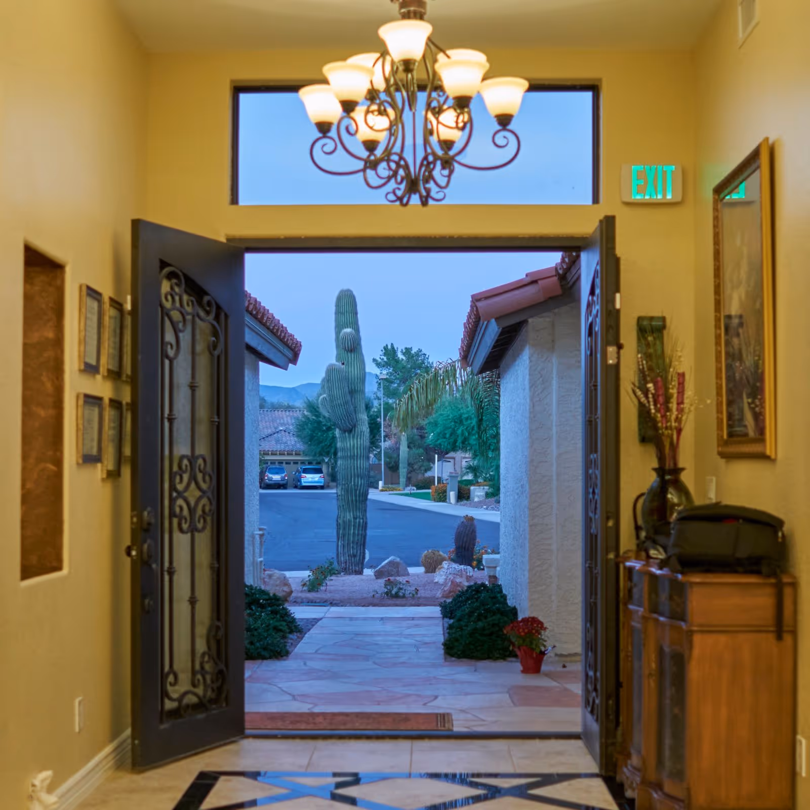 Foyer with an open wrought-iron double door and chandelier looking out onto a walkway and a large saguaro cactus outside.