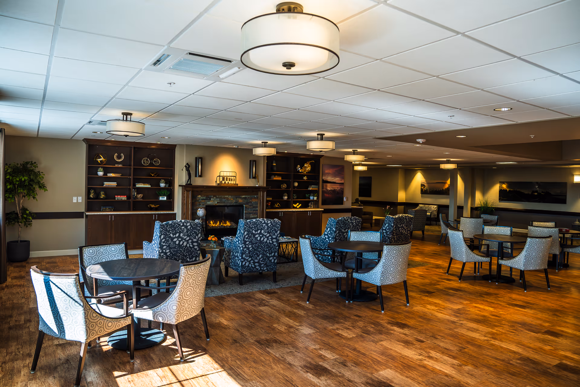 A spacious and well-lit common area with multiple round tables surrounded by patterned chairs. There are several blue armchairs arranged around a fireplace with a stone surround and wooden mantel. Built-in wooden shelves flank the fireplace, decorated with various ornaments and plants. The room has a wooden floor, ceiling lights, and neutral-colored walls with framed artwork.