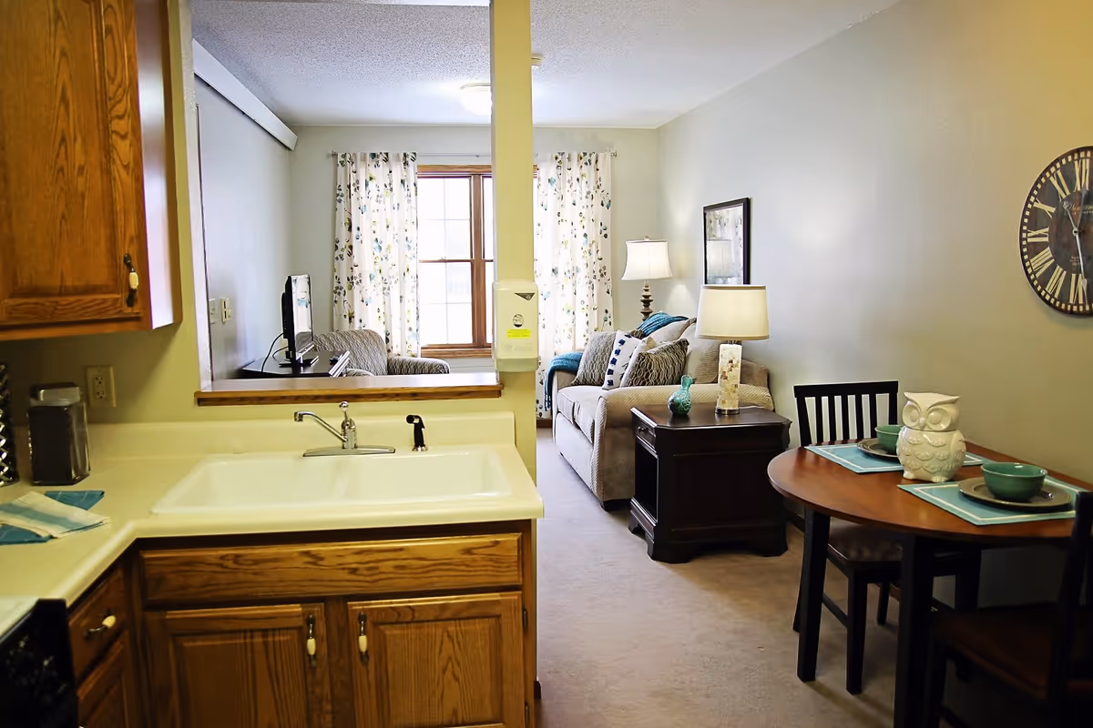 View of a small apartment interior showing a kitchen area with a sink and wooden cabinets in the foreground, a dining table set for two with an owl decoration on it, and a living room area with a sofa, side table with lamps, and a wall clock. A window with floral curtains is visible in the background.