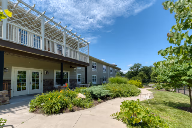 Outdoor view of Cedarhurst of Columbia showing a two-story building with a covered patio and balcony. The area features a paved walkway surrounded by green shrubs, plants, and trees under a partly cloudy blue sky. There are benches along the path for seating.