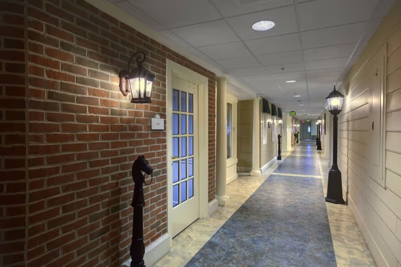 Indoor hallway with a brick wall on the left and a beige paneled wall on the right, featuring decorative horse head posts and lantern-style wall and floor lamps, leading to multiple doors and a distant exit sign.
