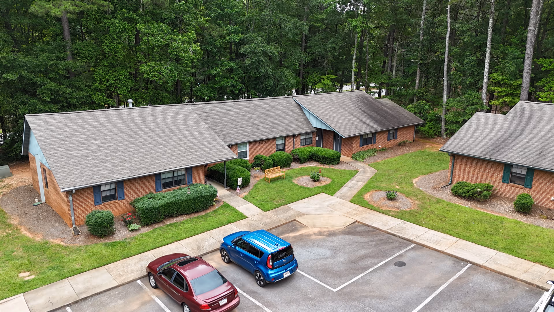 Aerial view of a senior living facility named Shenandoah Villas showing single-story brick buildings with gray shingled roofs surrounded by green grass and trees. There are two cars parked in the parking lot in front of the buildings, and small landscaped areas with bushes and a bench near the entrances.