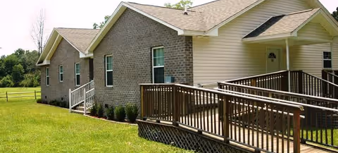 Exterior view of a single-story residential care facility building with a brick and siding facade, multiple windows, a wooden ramp with railings leading to the entrance, and a grassy lawn surrounding the building.