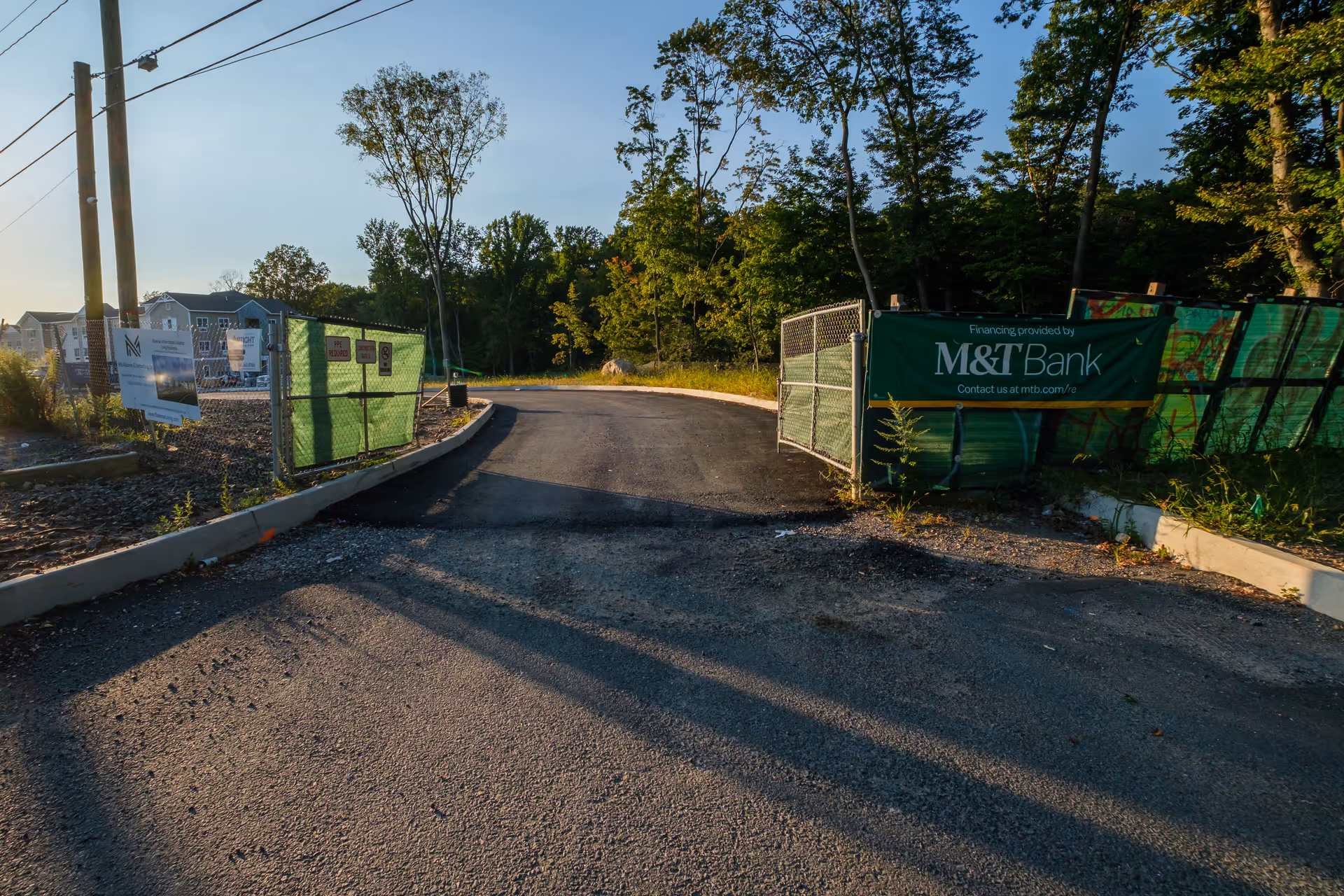 A paved road leading into a fenced construction site with green mesh fencing on both sides. There are signs on the fences, including one from M&T Bank indicating financing. Trees and houses are visible in the background under a clear sky.