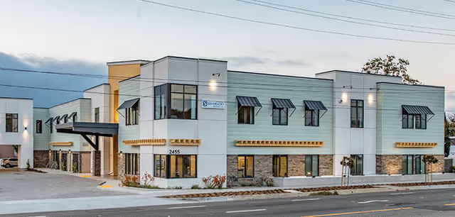 Two-story modern memory care building with pale green and white siding, multiple windows and awnings along a street at dusk.