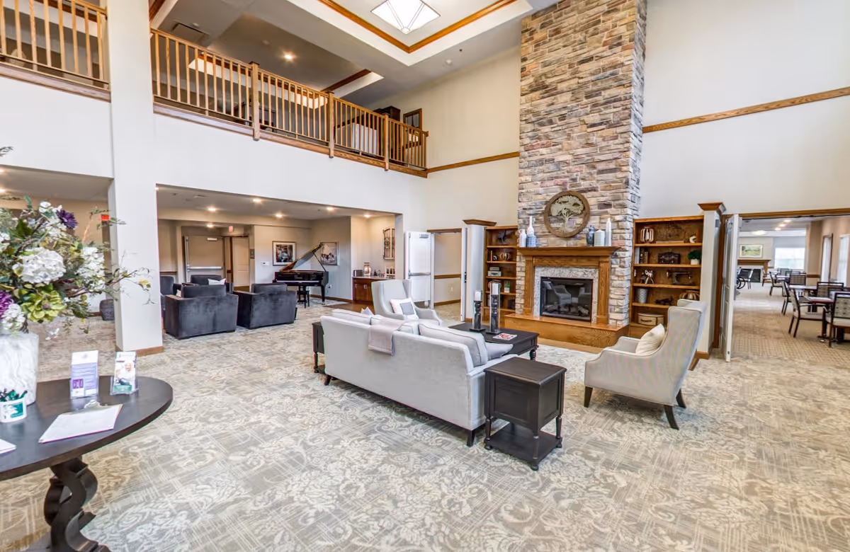 Spacious senior living facility common area with high ceilings, a stone fireplace, comfortable seating including sofas and armchairs, a grand piano in the background, and a balcony overlooking the space. There is a patterned carpet and a table with flowers and informational brochures near the entrance.