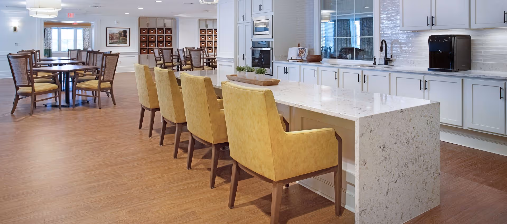 Bright communal dining area with a marble-topped island, yellow upholstered chairs, kitchenette cabinets, and round dining tables in the background.