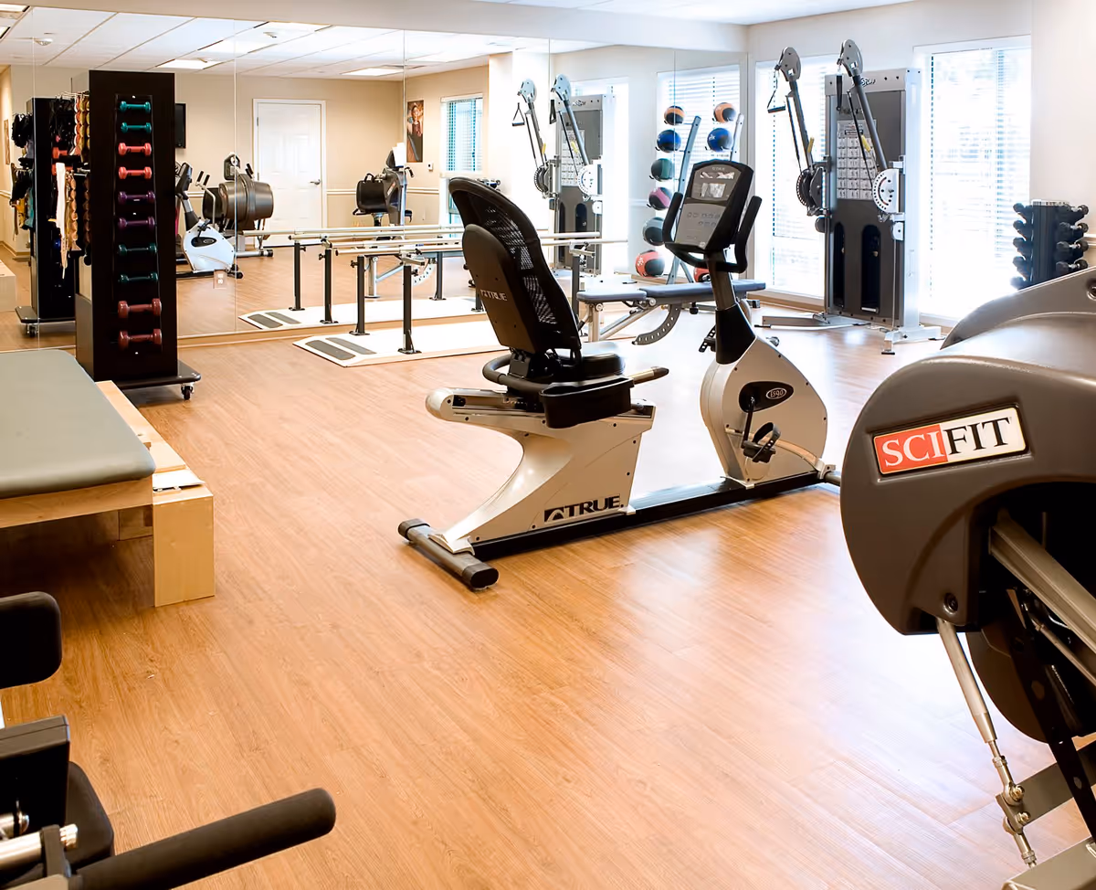 A bright fitness room with wooden flooring featuring various exercise equipment including a recumbent stationary bike, weight racks with dumbbells and medicine balls, parallel bars for walking exercises, and a padded bench. Large mirrors cover one wall and windows allow natural light to fill the space.