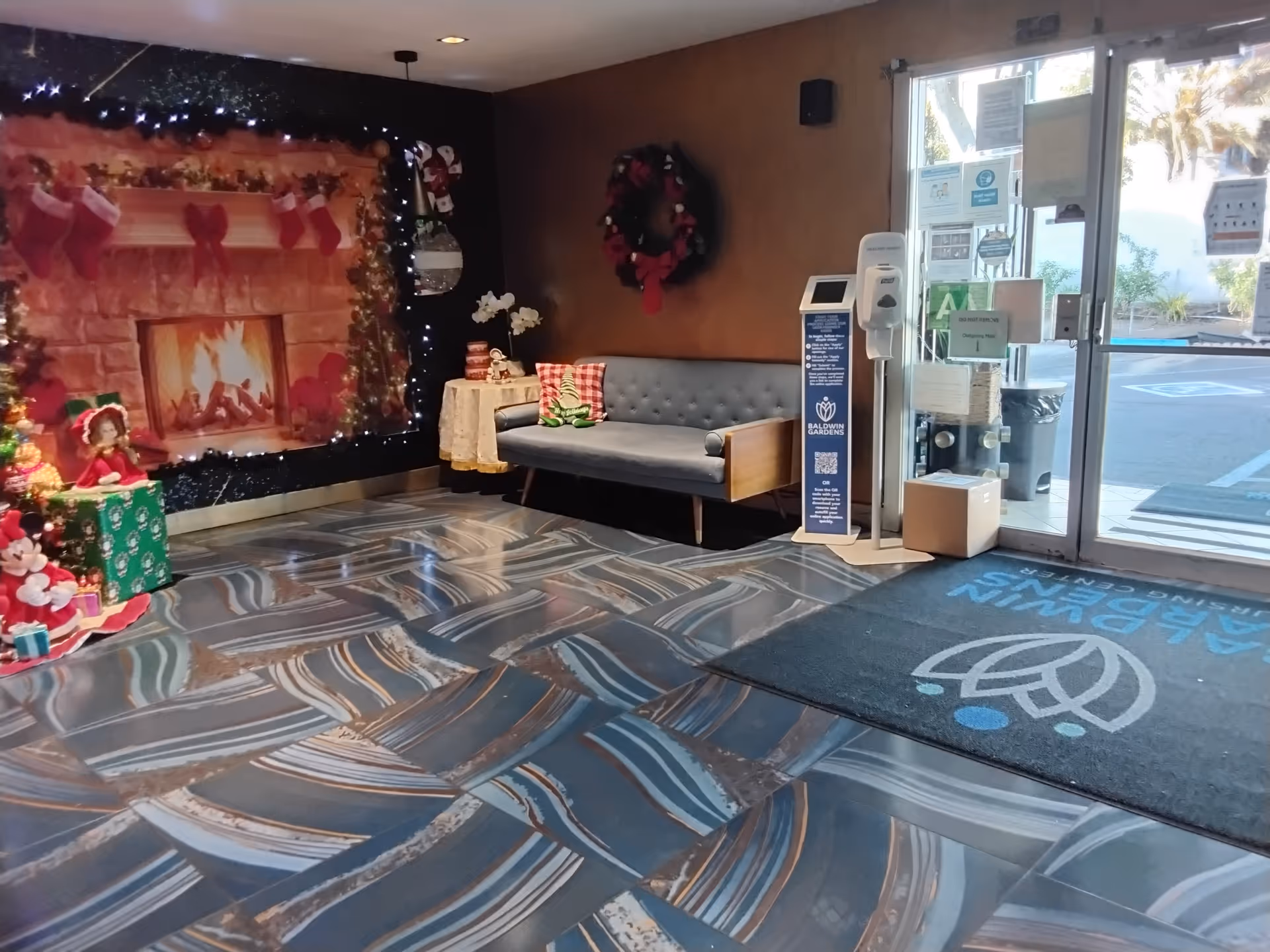 Lobby area of Baldwin Gardens Nursing Center decorated for Christmas with a large festive backdrop featuring a fireplace and stockings, a small Christmas tree with presents, a gray cushioned bench with holiday pillows, a wreath on the wall, and a glass entrance door with various notices and a hand sanitizer station nearby.