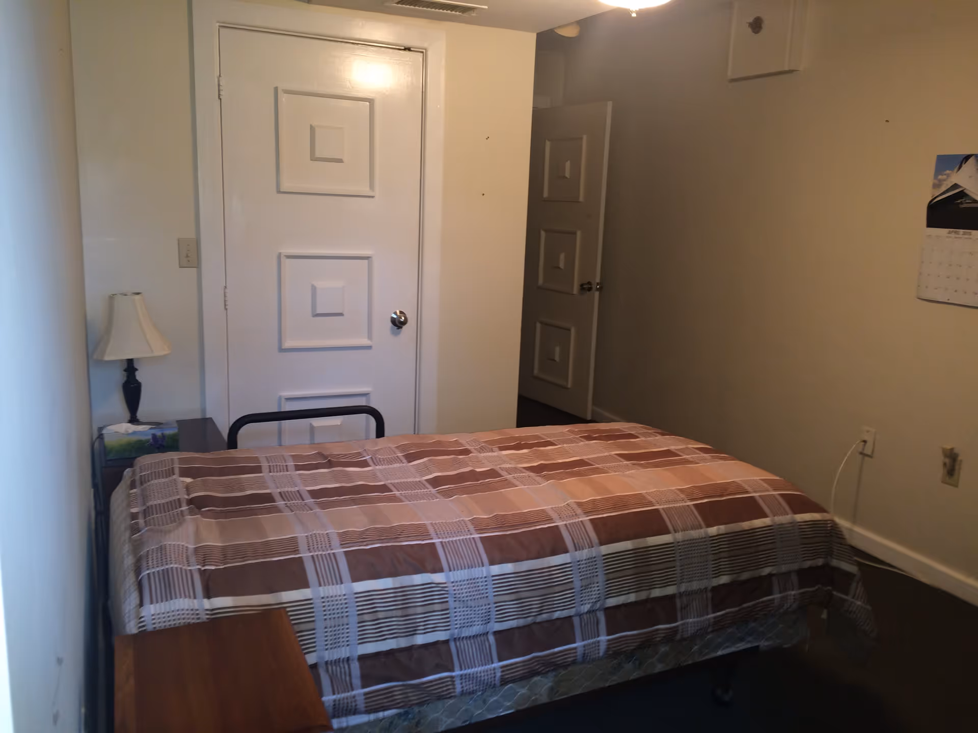 Small bedroom with a single bed covered in a brown plaid comforter, a nightstand with a lamp, and two white doors.