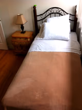 A single bed with a brown blanket, white sheets, and two white pillows. Next to the bed is a wooden nightstand with a decorative design and a table lamp with a beige shade. The room has wooden flooring and a white wall.