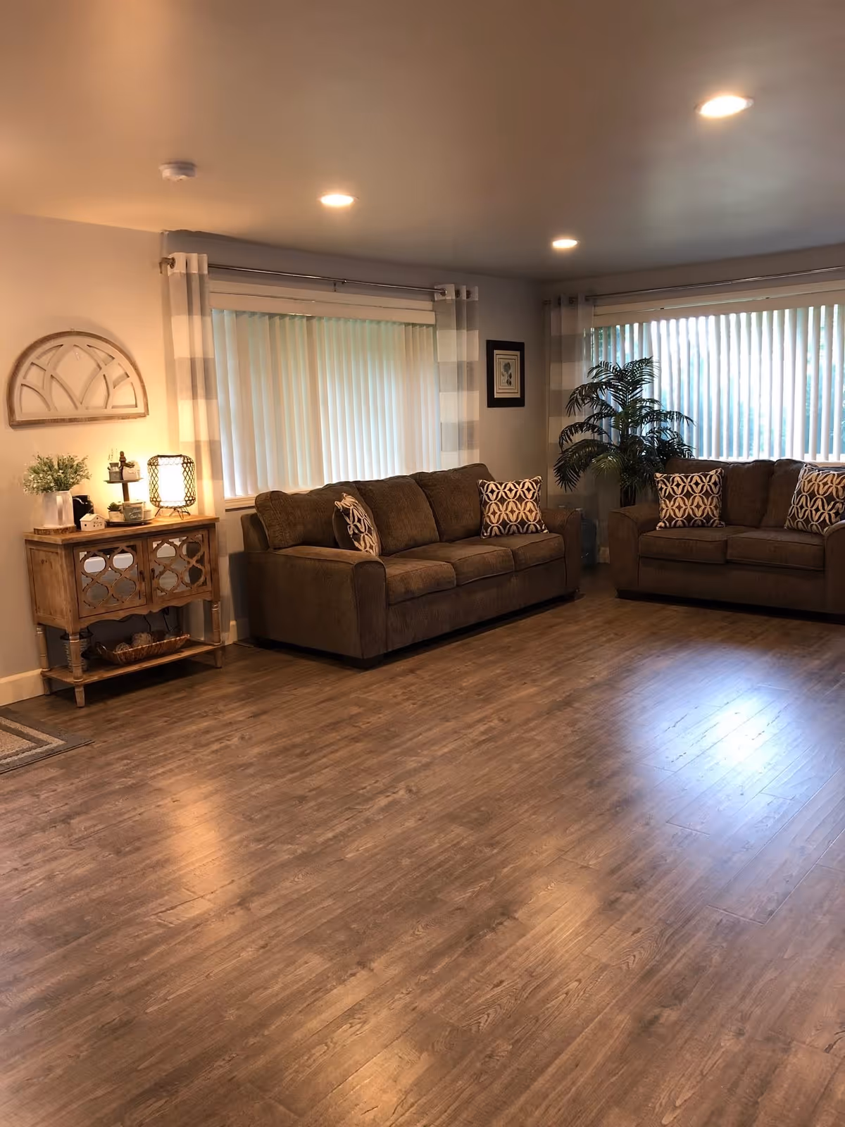 A cozy living room with two brown sofas adorned with patterned cushions, large windows with vertical blinds and striped curtains, a wooden side table with decorative items and a lamp, a potted plant in the corner, and wood flooring.