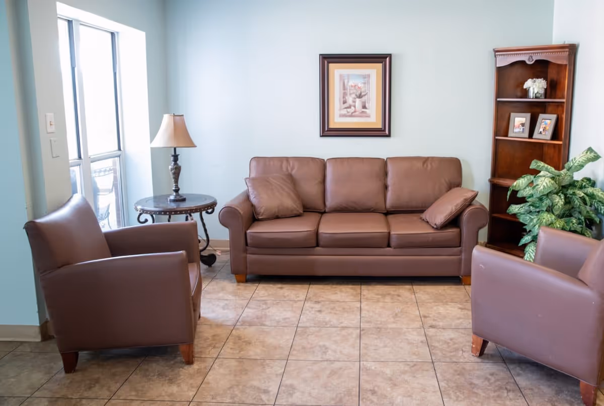 Small seating area with a brown leather sofa and armchairs, side table with lamp, bookshelf, framed artwork, and potted plant.
