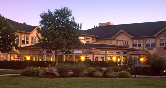 Exterior view of a senior living facility building at dusk with illuminated windows, landscaped garden with bushes and trees, and benches in the foreground.
