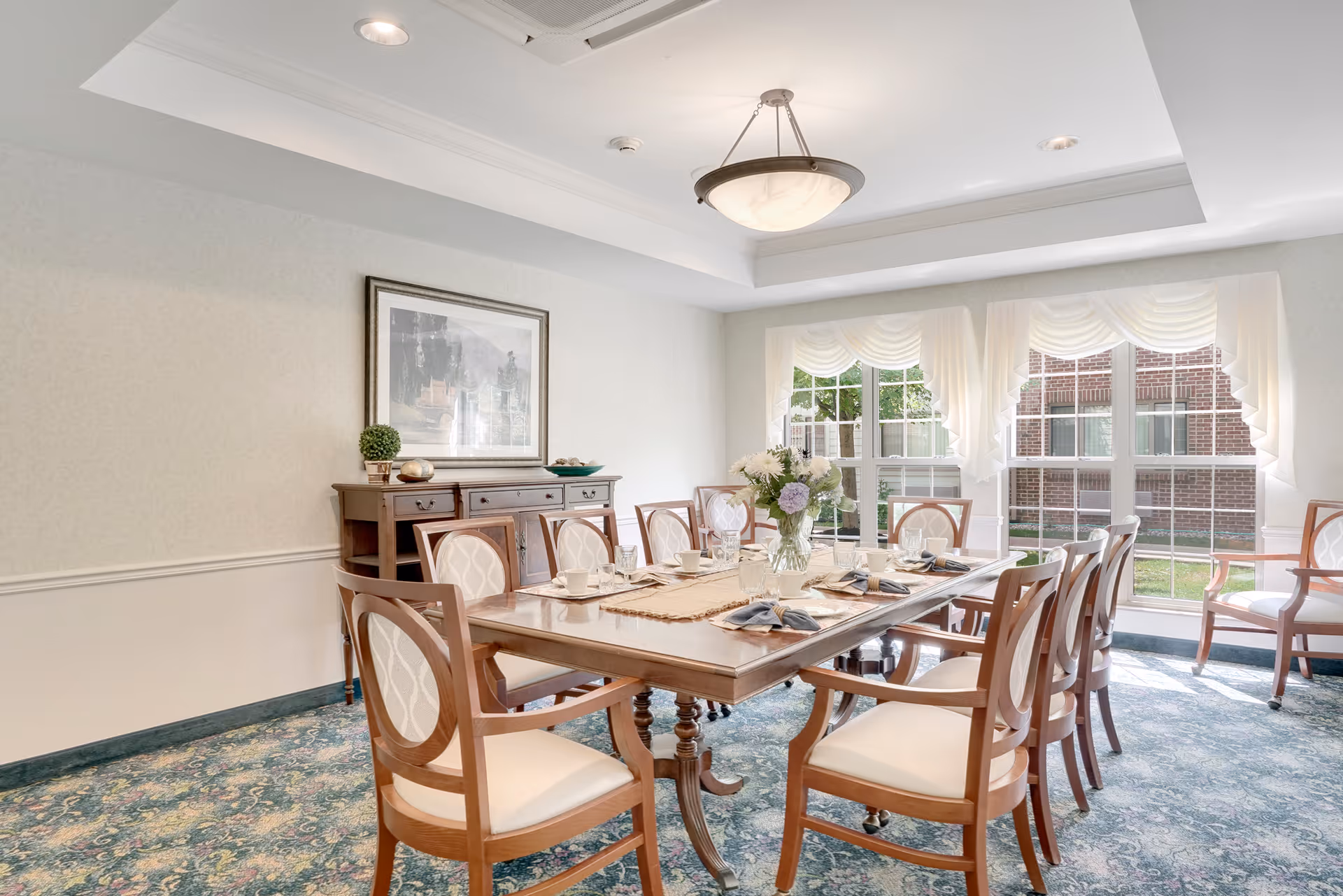 A bright dining room with a large wooden dining table set for eight people. The table has a centerpiece of flowers, cups, glasses, and napkins. The room features large windows with white curtains allowing natural light to fill the space. There is a wooden sideboard against the wall with decorative items and a framed picture above it. The floor is carpeted with a patterned design.