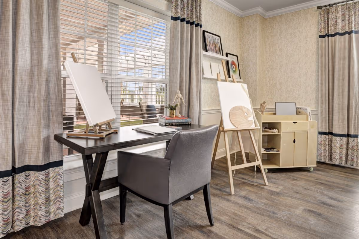 A bright art room with a large window covered by blinds and curtains. There is a dark wooden table with a gray upholstered chair, two blank canvases on easels, a small stack of books, and art supplies. The room has light-colored wallpaper, wooden flooring, and a small rolling cabinet with shelves and drawers.
