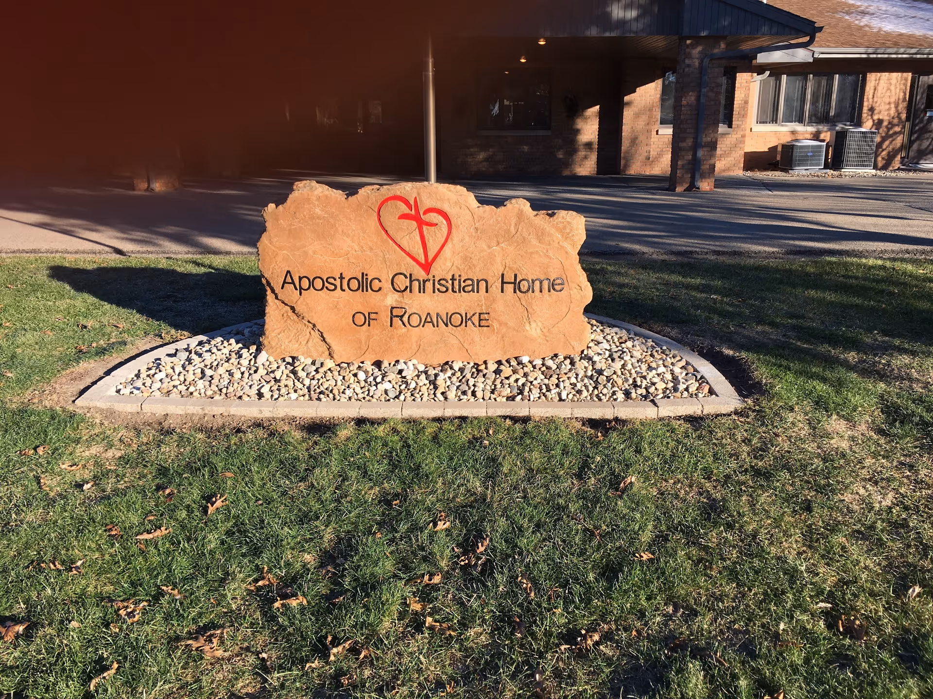 Stone sign that reads "Apostolic Christian Home of Roanoke" set on rocks and grass in front of the facility entrance.