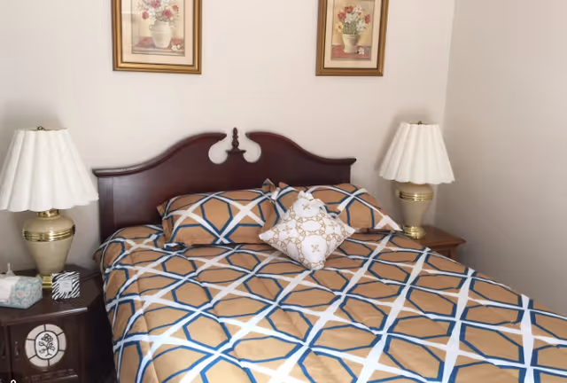 A bedroom with a large bed featuring a geometric patterned bedspread in beige, blue, and white. The bed has a dark wooden headboard and two matching pillows along with a decorative white pillow in the center. On each side of the bed are wooden nightstands with beige lamps with white lampshades. Above the bed are two framed floral paintings on a light-colored wall.