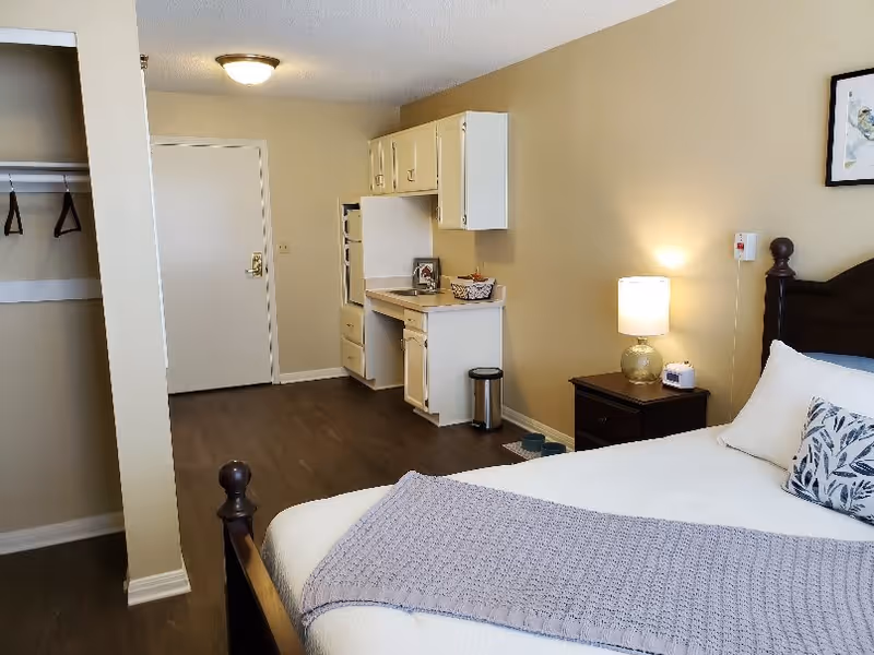A senior living facility room with a bed featuring white linens and a lavender throw blanket. There is a bedside table with a lamp and a small device on it. The room has a kitchenette area with white cabinets, a small countertop, and a trash can. The floor is wooden, and there is an open closet with hangers on the left side. The walls are painted beige, and a framed picture hangs above the bed.