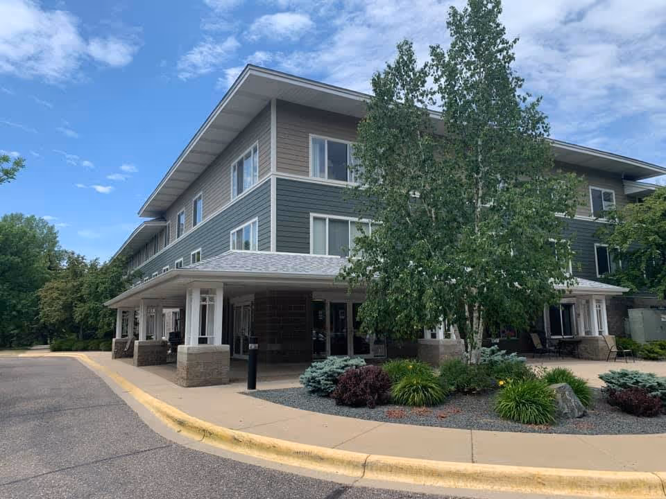 Exterior view of a three-story senior living facility building with a covered entrance, surrounded by trees and landscaped bushes under a partly cloudy sky.