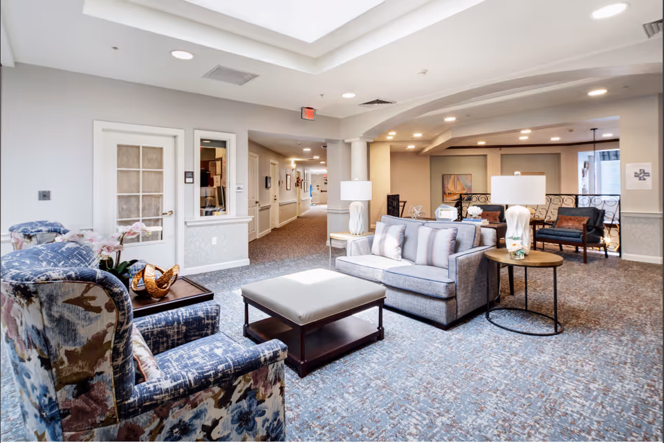 A bright and spacious senior living facility common area with patterned armchairs, a gray sofa with striped pillows, a cushioned ottoman, side tables with lamps, and decorative items. The area has carpeted floors, soft lighting, and a hallway leading to other rooms.