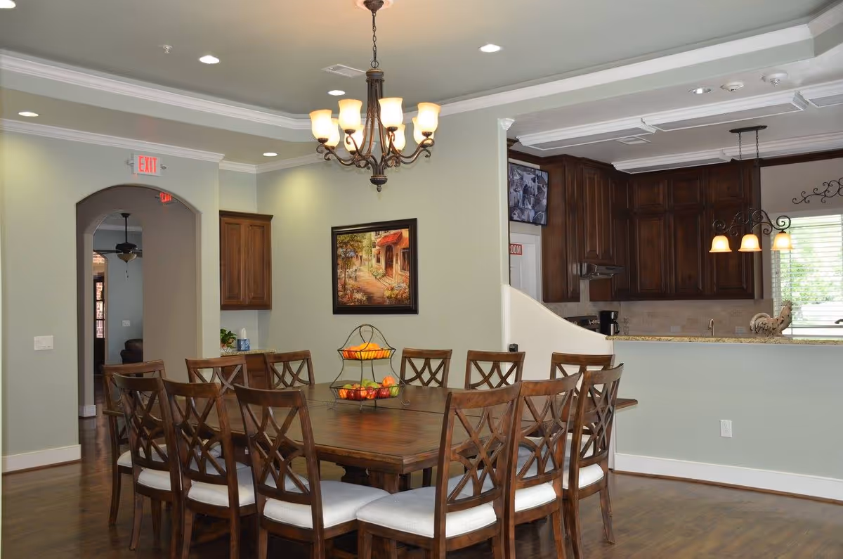 Spacious dining room with a large wooden table and chairs, a chandelier overhead, and an open view into a kitchen with dark cabinets.