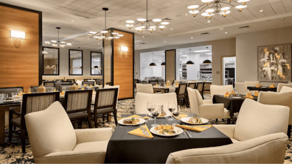 A spacious and elegant dining room with multiple tables set for a meal. The tables have black tablecloths, plates with food, wine glasses, and yellow napkins. The room features comfortable beige upholstered chairs, modern light fixtures on the ceiling, wood-paneled walls, and decorative artwork. The background shows an open kitchen area with pendant lights.
