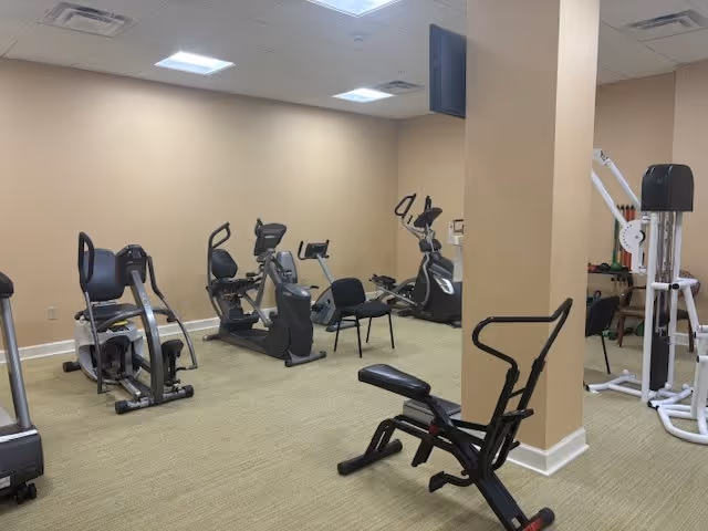 A senior living facility exercise room with various fitness equipment including stationary bikes, an elliptical machine, a weight machine, and a bench. The room has beige walls, a carpeted floor, and a mounted television on the wall.