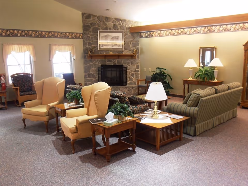 A cozy living room area in a retirement community featuring two beige wingback chairs, a striped sofa, a floral patterned loveseat, wooden side tables with lamps, and a stone fireplace with a framed picture above it. The room has carpeted flooring, windows with curtains, and decorative plants.