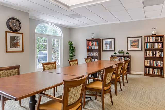 A well-lit room with a long wooden table surrounded by cushioned chairs. The room has beige walls with framed artwork and a clock. There are two tall bookshelves filled with books and a small cabinet with decorative plants. A large window with an arched top allows natural light to enter, and a potted plant is placed near the window.