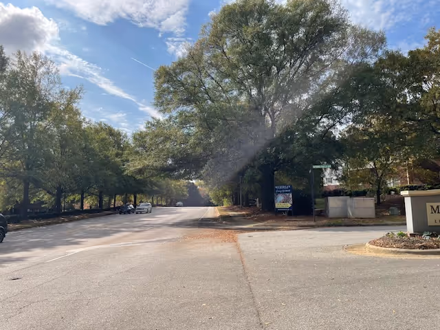 A wide paved road lined with large trees on both sides under a partly cloudy sky. There are a few parked cars along the left side of the road and a sign on the right side near the curb. The area appears to be part of a residential or community complex.