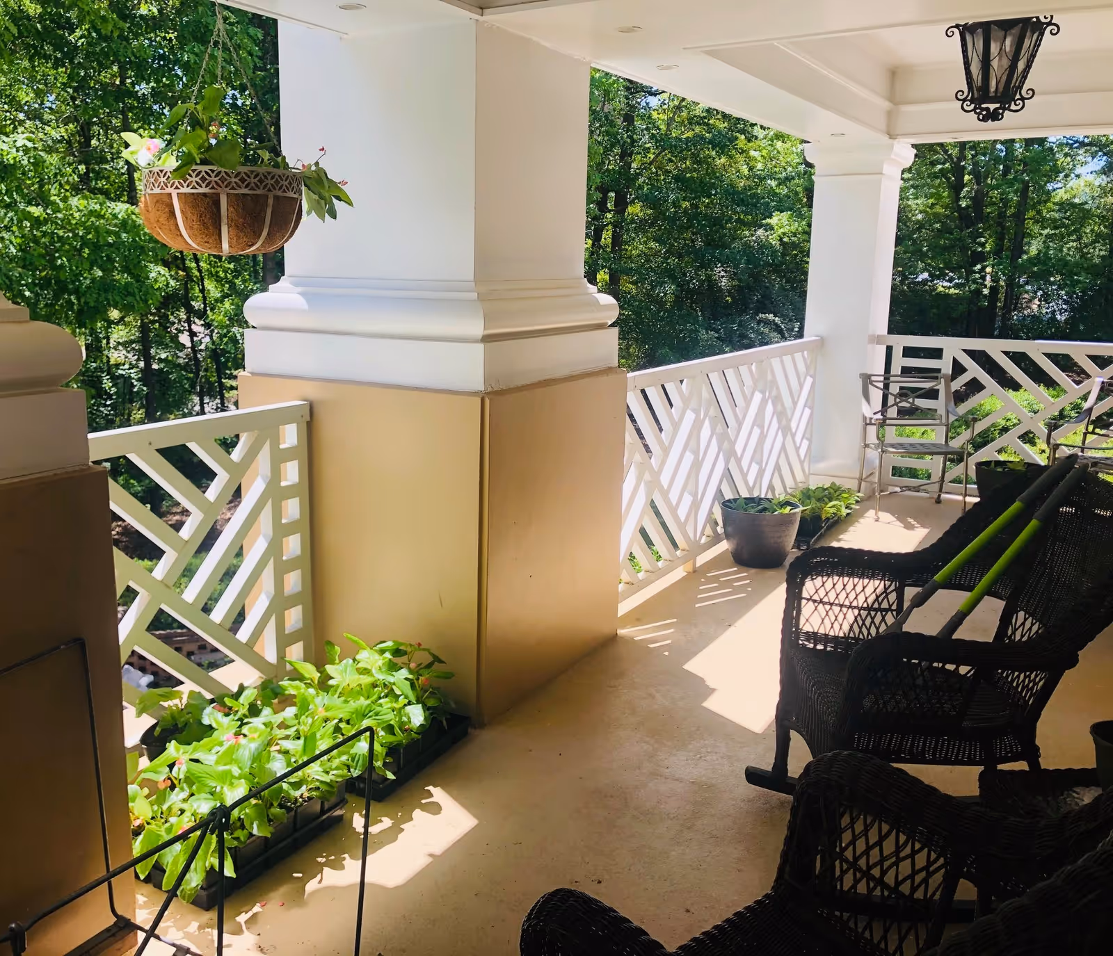 A covered outdoor balcony area with white railings and large white columns. The balcony has several black wicker chairs and a metal chair, with potted plants and hanging flower baskets. Trees and greenery are visible in the background.