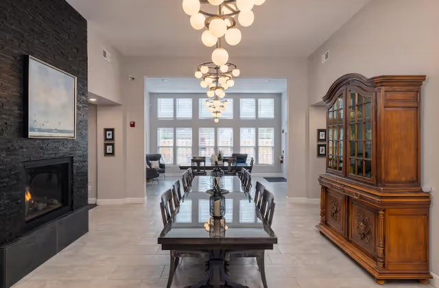 A bright communal dining area with a long wooden table and chairs, modern chandeliers, a fireplace on the left and a large wooden cabinet on the right.