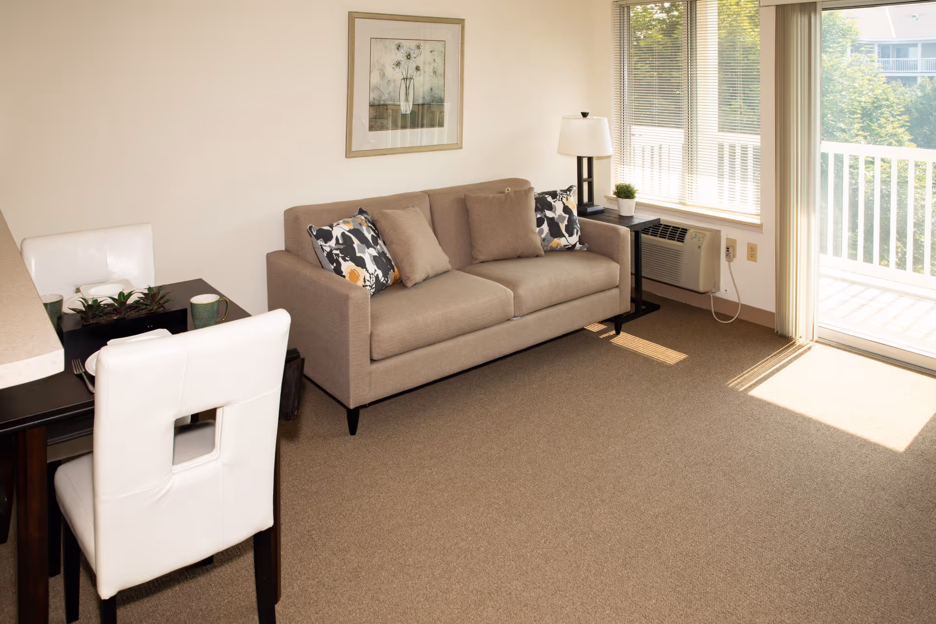 A cozy senior living room with a beige sofa adorned with patterned and plain cushions, a side table with a lamp and small plant, a dining table with white cushioned chairs, and large windows with blinds letting in natural light. A sliding glass door opens to a balcony with a white railing and greenery outside.