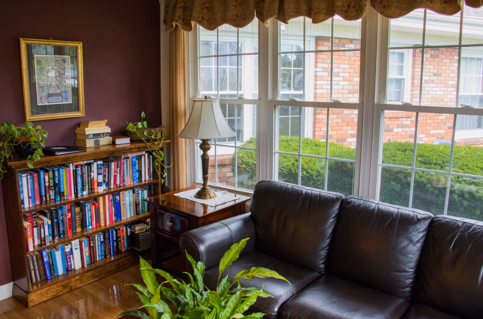 Cozy living room area with a dark leather couch, wooden side table with a lamp, a bookshelf filled with books, potted plants, and large windows showing a view of green bushes and a brick building outside.