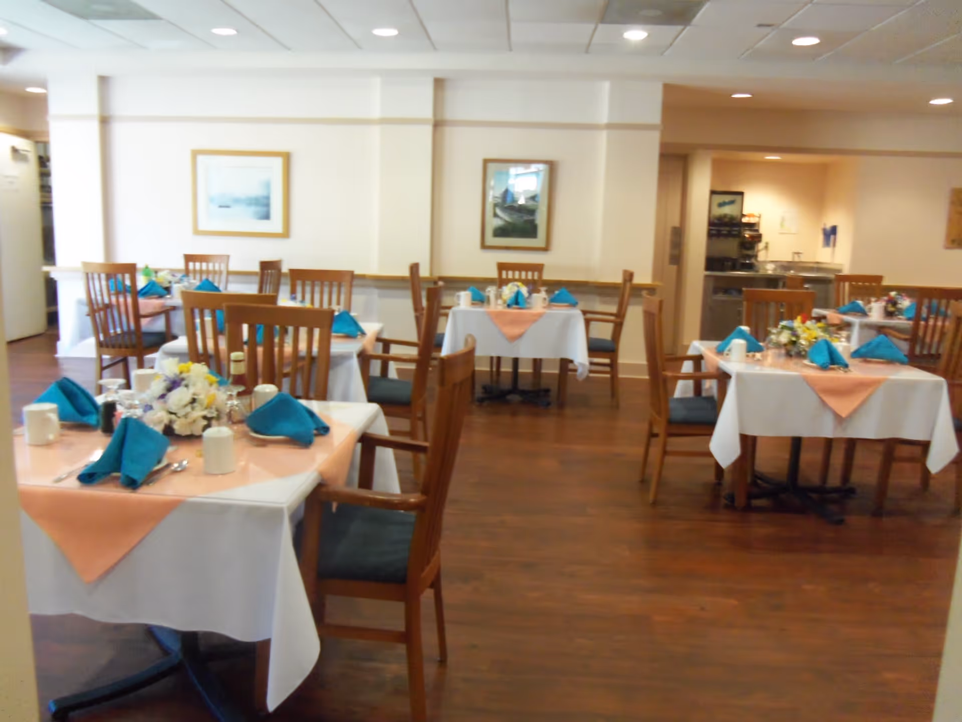 A dining room in a senior living community with several tables covered in white tablecloths and peach-colored overlays. Each table is set with blue folded napkins, white mugs, silverware, and floral centerpieces. Wooden chairs with dark cushions surround the tables. The room has wooden flooring, light-colored walls, framed artwork, and a ceiling with recessed lighting.