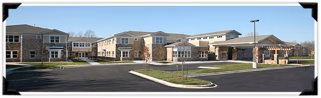 Wide exterior view of a large, modern senior living facility building with stone and beige siding, multiple windows, and a covered entrance. The building is surrounded by a paved driveway and parking area with some small trees and landscaping under a clear blue sky.
