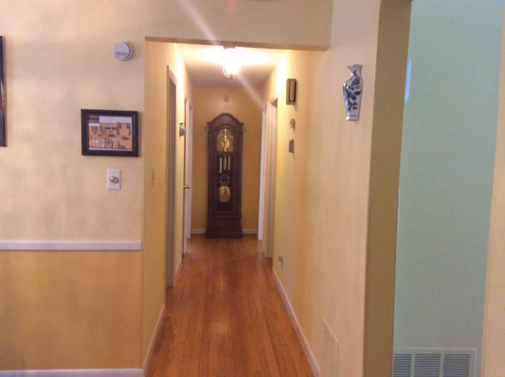 A narrow interior hallway with hardwood floors and yellow walls leading to a grandfather clock at the end.