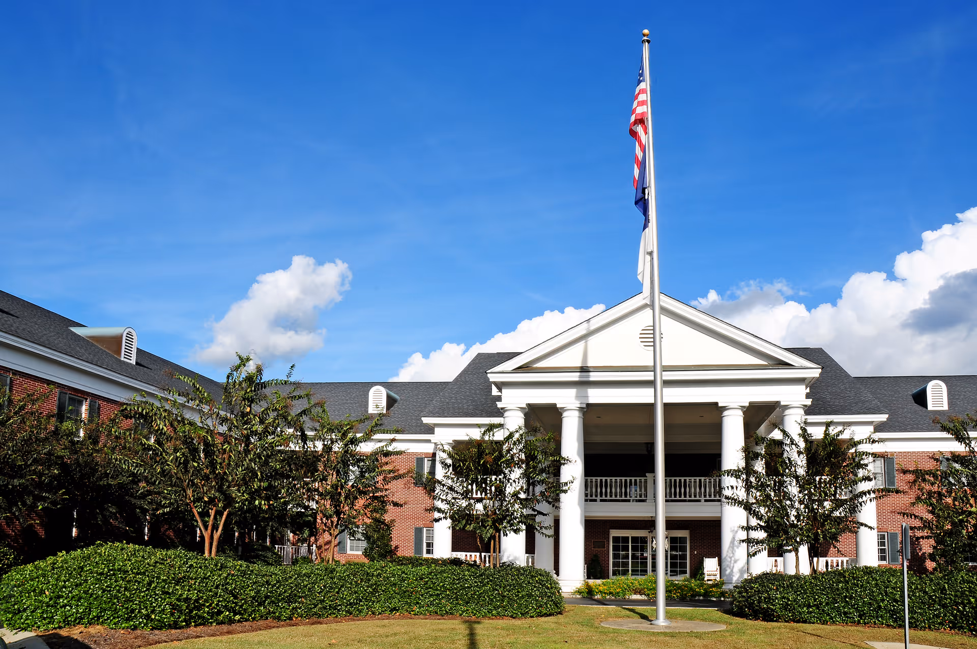 Front exterior view of a large brick building with white columns and a covered entrance. There is a flagpole with an American flag in front of the building, surrounded by green bushes and trees under a blue sky with some clouds.